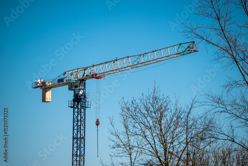 Tall tower crane with a lattice jib extends horizontally against a clear blue sky. A hook hangs from the jib's end by a cable. 