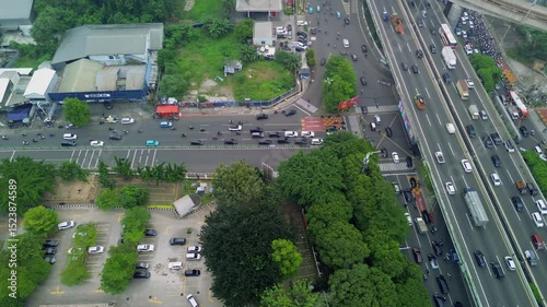 This aerial footage showcases a bustling highway in Jakarta, Indonesia. The scene includes cars, trucks, motorcycles, buildings, trees, and a parking lot. The camera slowly pans across the landscape.