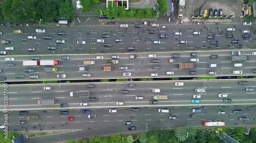 This aerial footage showcases heavy traffic flow on a wide highway. The scene features cars, trucks, buses, and motorcycles moving along multiple lanes, surrounded by green trees.
