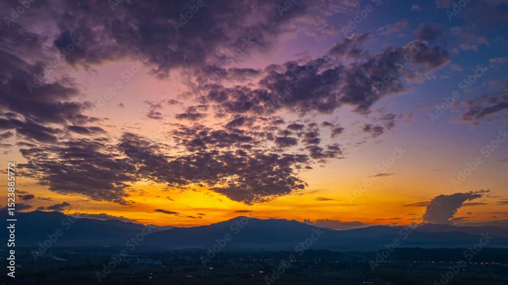 Obraz premium A breathtaking aerial panoramic view of a vibrant sunset illuminating the sky above a lush green hilltop with a small structure, surrounded by scattered clouds and distant landscapes.