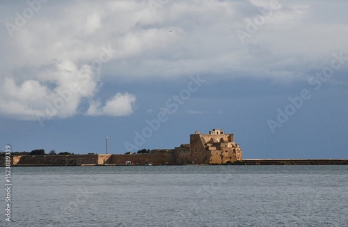Sunlight falls on ruined stone fortifications on a harbour breakwater. The sky is overcast and the water is grey. The fort and defensive wall are of sandstone.