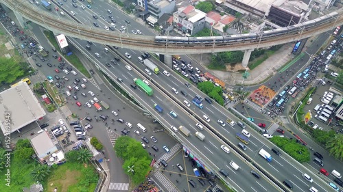 An aerial shot shows heavy traffic on a Jakarta highway. A train crosses overhead on a railway bridge. Buildings and greenery surround the busy road.