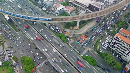 This aerial footage shows heavy traffic in Jakarta, Indonesia. Cars, buses, and motorcycles navigate the roads, with a train crossing over on a bridge. The shot is taken from above.
