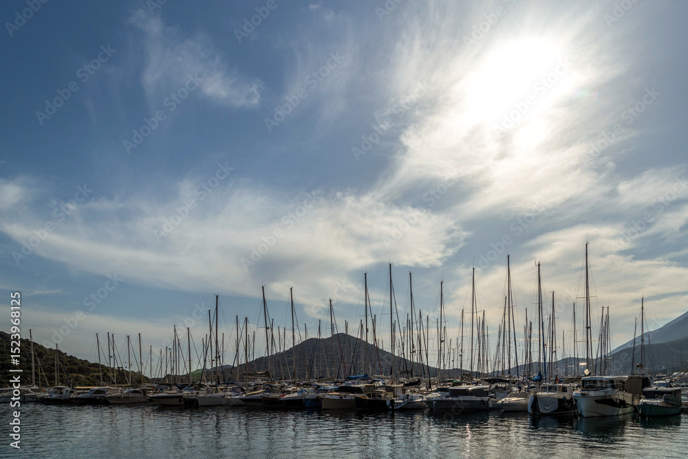 Obraz premium Many sailboats with tall masts docked at marina pier, backlit by sun. Reflections on water, blue sky, green hills in background. Kas, Mediterranean, Turkey.