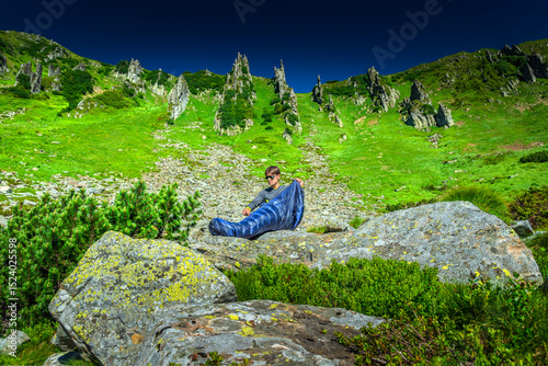 Man unzipping a sleeping bag while sitting on rocky terrain in a lush green alpine valley under a clear blue sky, during a hiking and camping morning routine. Outdoor adventure concept
