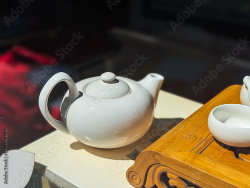 White ceramic teapot on light surface. Round teapot with curved handle and short spout next to bamboo tea tray.