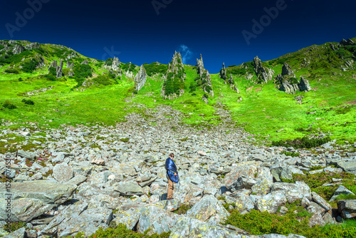 Young man wrapped in a sleeping bag standing on a rocky slope in the mountains under a deep blue sky. Summer hiking, camping, and outdoor adventure in wild alpine landscape