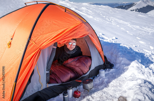 Man resting in sleeping bag inside orange tent on snowy mountain. Camping stove, thermos and cup on snow in foreground. Winter camping, outdoor survival, mountain adventure