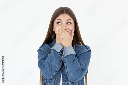 Portrait of a shocked young girl in jeans coat looking aside with mouth covered isolated over white background