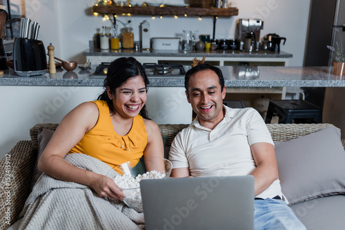 young Latin couple watching a movie on sofa at home in Mexico Latin America, hispanic people eating popcorn at home