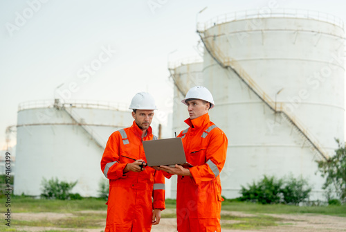 Side view of two engineers or gas factory technicians use laptop to discuss together about work and stand in front of gas tank in workplace.