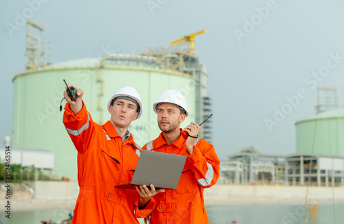 One gas factory worker use walkie talkie point to his right side and discuss with coworker to solve the problem and stay in front of gas factory tank near the bay.