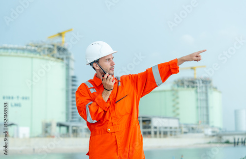 Petrochemical or gas factory worker talk with walkie talkie and point to his left side in front of tank in factory.