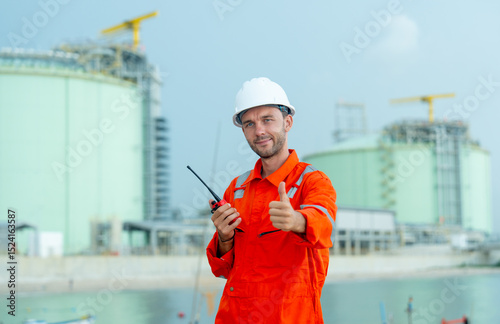 Petrochemical or gas factory worker hold walkie talkie and show thumbs up to camera and stand in front of tank in factory.