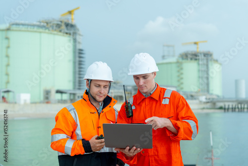 Two petrochemical or gas factory workers men stand in front of factory tank near the bay and work together using laptop.