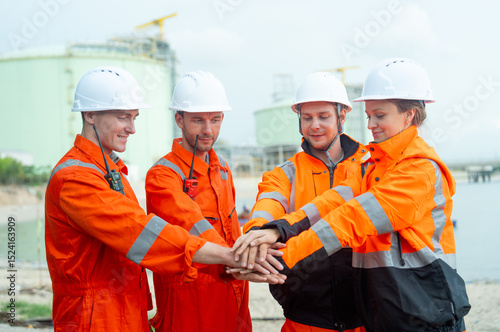 Group of petrochemical workers or technicians stand and hold hands together in front of factory tank.