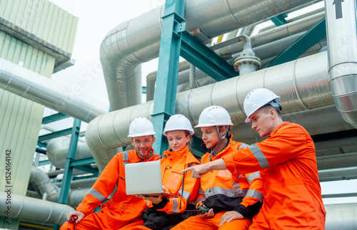 Side view group of petrochemical or gas factory workers or engineers discuss about work using laptop and sit near pipeline in factory workplace.