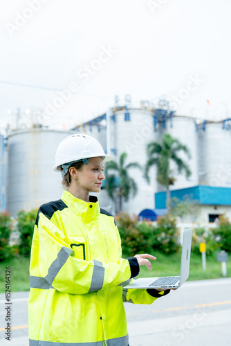Vertical image of petrochemical or gas factory worker hold laptop for working and look to her left side in front of the factory.