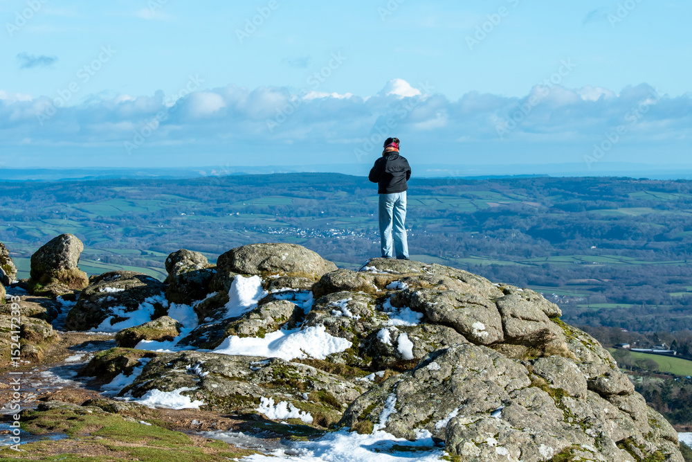 Obraz premium Person standing on a tor at Dartmoor National park, looking out at the vast hills and valleys.