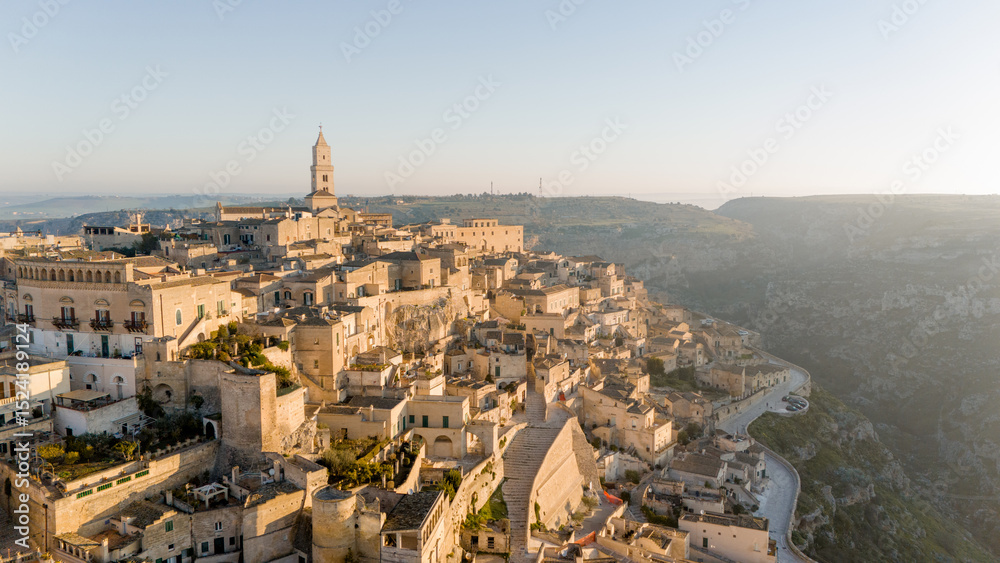 Fototapeta premium Aerial view of Matera, Italy, bathed in golden morning light. The iconic stone dwellings and cathedral tower rise above the canyon, showcasing this UNESCO-listed historic wonder