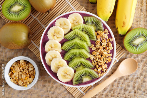 A vibrant acai bowl, adorned with sliced kiwi and banana, sits atop a burlap surface, next to a small bowl of granola