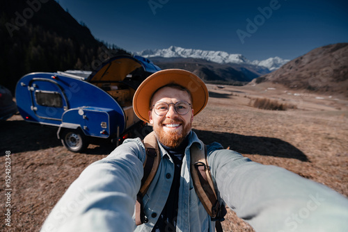 Young happy man hipster traveler making selfie background camper car on mountain. Concept one life adventure trip, vacation. photo