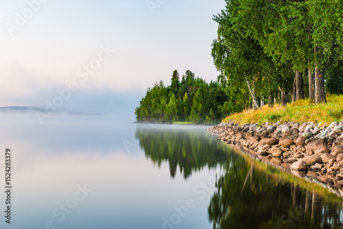 Fototapeta Naklejka Na Ścianę i Meble -  Misty morning on the still lake in Sweden with lush greenery lining the shore