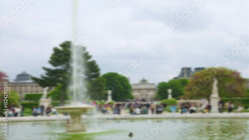 People walk and relax around the Grand bassin rond with fountain in Jardin des Tuileries with overlooking on central alley and Arc de Triomphe du Carrousel in 1st arrondissement. Out of focus. Paris.
