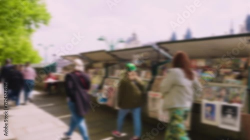 People stroll along the Quai de la Megisserie embankment in spring past green metal kiosks with souvenirs and posters. Out of focus. Paris, France.