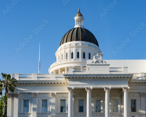 Detailed close-up of the California State Capitol building, highlighting its ornate dome, white columns, and classic architecture under a bright blue sky, with palm trees and a flag adding to the Sacr