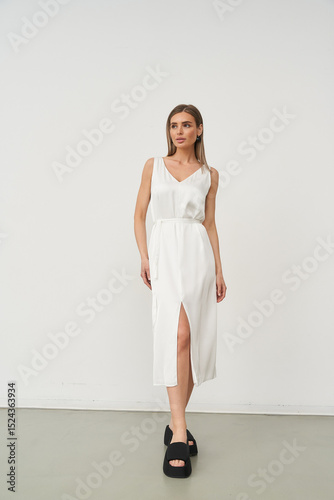 Elegant woman in fitted white dress with a front slit, posing confidently, wearing black platform sandals. She stands against a neutral backdrop, exuding sophistication and style