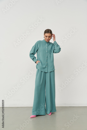Woman in a teal button-up shirt and wide-leg pants, with hands in pockets. She wears pink shoes and poses confidently, looking down slightly, with hair neatly styled back