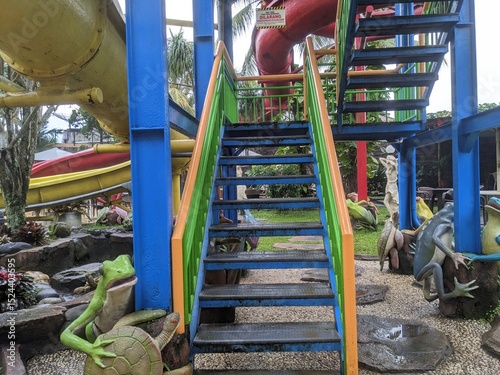 Colorful waterpark staircase with slides, frog statues, and playful decorations in a tropical outdoor setting after rain.
