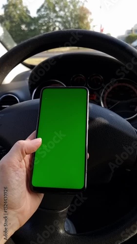 A hand holds a smartphone with a green screen vertically in a car against the background of a busy road. Vertical video.
