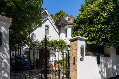 Fényképezés London, UK, 5 May 2025,  A stately white house with architectural details, partially obscured by trees and viewed through ornate black iron gates