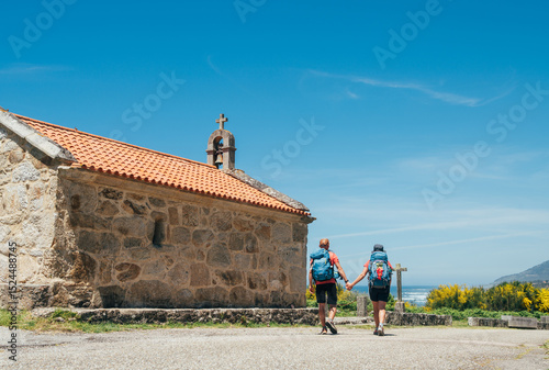 Fotografie Couple of pilgrims with backpacks walking hand in hand next to old church on Camino Portuguese Way, inspiring walk on famous Camino de Santiago