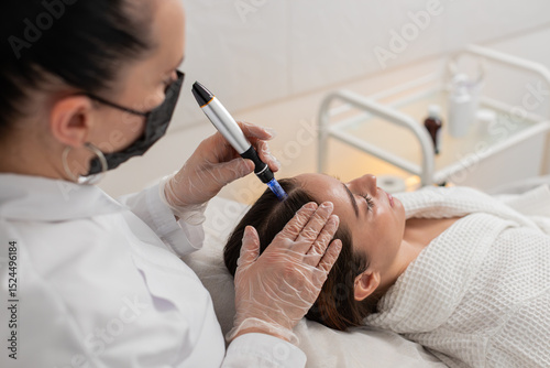 Caucasian girl undergoes scalp treatment to stimulate hair regrowth using microneedling dermapen and vitamin injections. Procedure performed by trichologist in cosmetology clinic for hair restoration.