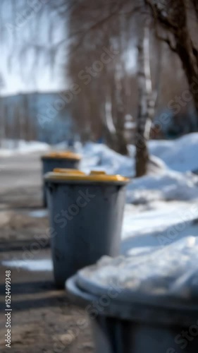 Row of gray trash bins along a wintry urban street with melting snow and bare trees in soft daylight