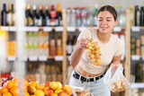 In self-service minimarket, girl customer takes out grape from showcase, examines fruits and buys goods. She picks ripe fruits berries from large box in stores storage
