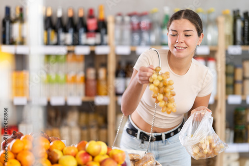 Wallpaper Mural In self-service minimarket, girl customer takes out grape from showcase, examines fruits and buys goods. She picks ripe fruits berries from large box in stores storage Torontodigital.ca