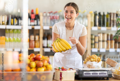 While working at grocery store. Young woman in white uniform works as salesperson, sells bananas, serves packaged fruits. Trading floor of store, assortment of goods on shelves and showcases.