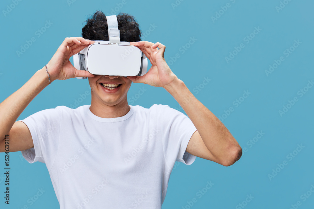 Naklejka premium Young man wearing virtual reality headset enjoying immersive digital experience, smiling and relaxed against blue background in casual white t-shirt.
