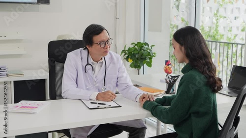 Middle-aged Asian male doctor checking medical results of female patient on tablet in clinic, discussing medical issues with patient during professional healthcare consultation.