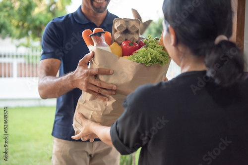 Black male Delivery person hands over a brown paper bag filled with fresh vegetables and fruits to a smiling customer in a residential area.