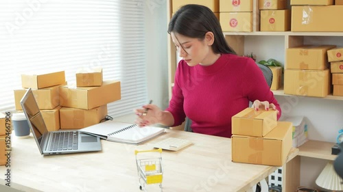 SME entrepreneur holding laptop focusing on online sales and customer service on digital tablet while packing boxes at home. Using social media for marketing. Online shopping.