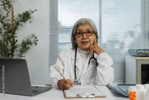 Experienced Asian female doctor wearing a lab coat and stethoscope, diligently writing notes on a clipboard while seated at her desk in a professional medical office setting photo
