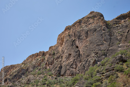 Scenic view of a rugged rocky cliff under a bright blue sky in a dry mountainous region. Natural textures, arid landscape, and distant hills create a stunning outdoor wilderness scene.