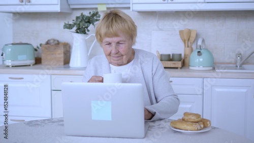 Senior woman sipping coffee while using a laptop in a bright kitchen, savoring her morning routine with delicious breakfast pastries and enjoying a moment of digital connection