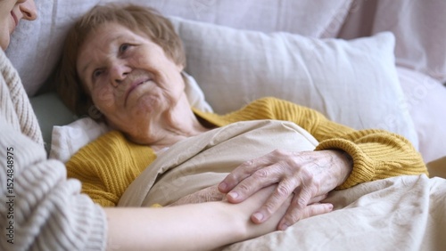 Photos Young nurse holding hands with elderly patient lying in bed, providing comfort a