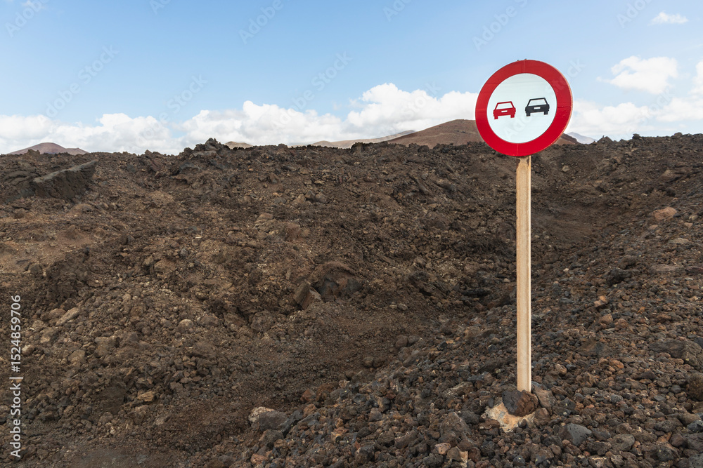 A road sign stands on rugged, dark volcanic soil in Lanzarote, Canary Islands, Spain. The sign features a red circle with symbols of two cars, indicating a potential traffic restriction. A road sign stands on rugged, dark volcanic soil in Lanzarote, Canary Islands, Spain. The sign features a red circle with symbols of two cars, indicating a potential traffic restriction.